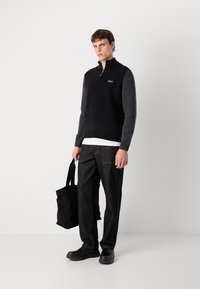 Sweater escuro com fecho éclair e mangas cinzas, usado sobre uma camisa branca. O modelo segura uma bolsa tote preta e veste calças escuras e sapatos robustos.