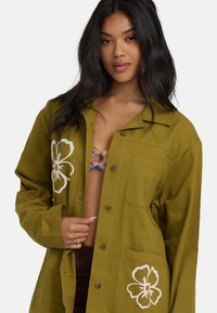 Veste chemise vert olive avec broderies florales, fermeture à boutons, col pointu, deux poches avant et tissu texturé.