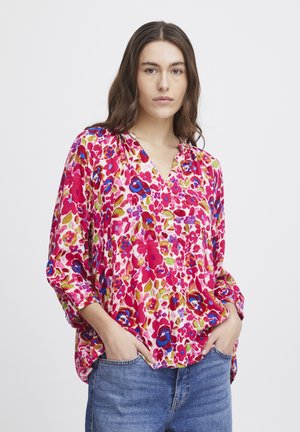 La blouse florale présente un fond rose vif avec des motifs de fleurs bleues et vertes. Elle a un col en V et des manches trois-quarts, fabriquée dans un tissu doux.