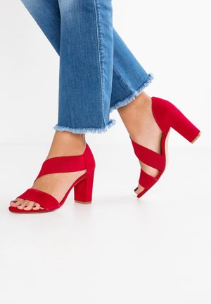 Fødder iført blå jeans med frynset kant og røde blokhælsandaler med krydsede elastiske stropper på hvidt gulv.