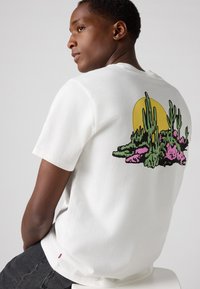 Hvit bomulls t-skjorte med grafisk design av kaktuser og blomster i rosa og grønn, plassert mot en gul solbakgrunn på baksiden.