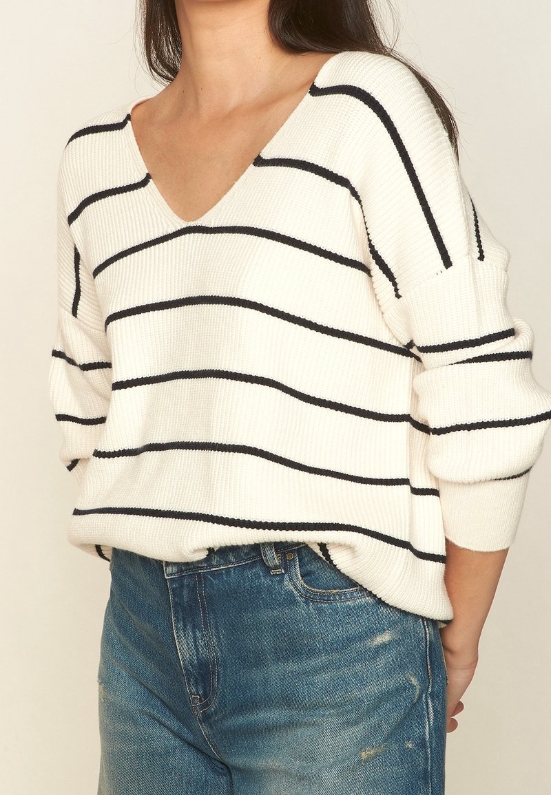 Femme portant un pull en maille blanc avec des rayures horizontales noires et un jean bleu déchiré, debout devant un fond beige uni.
