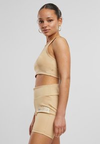 Beige treningssett som inkluderer en cropped tank topp og høyhalsede shorts. Stoffet er glatt med minimale sømmer og en fremtredende logo på shortsene.