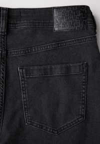 Svarta denimjeans med en texturerad svart midjebandsetikett med geometriskt mönster, en bakficka och synliga sömnadsdetaljer.