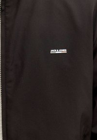 Λογότυπο "JACK & JONES" με χρωματιστή λωρίδα από κάτω σε μαύρο ύφασμα, που δείχνει μέρος ενός σκοτεινού ενδύματος με λεπτομέρεια ραφής.