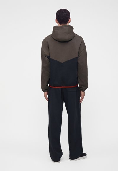 Homme debout, dos visible, portant une veste à capuche bicolore vert foncé et noir, un pantalon large foncé et des baskets noires à semelles blanches.