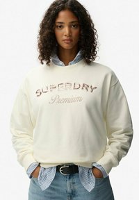 Sudadera crema con "SUPERDRY" bordado en rosa y "Premium" debajo. Manga larga, corte relajado, llevada sobre una camisa de rayas azul claro.