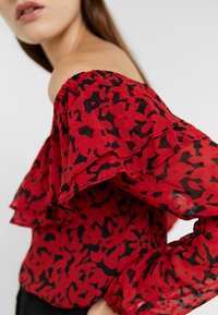 Blouse rouge à épaules dénudées avec bordure à volants, présentant un motif abstrait noir et une texture transparente. Design léger et ajusté qui met en valeur le décolleté.