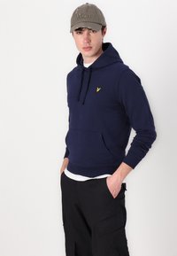 Felpa con cappuccio blu navy, dotata di tasca a canguro e logo giallo. Abbinata a pantaloni cargo neri. Il modello indossa un cappellino grigio con la scritta "Won" ricamata.