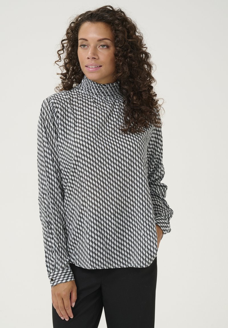 Blusa de patrones en blanco y negro con cuello de embudo, mangas largas y corte relajado. La tela presenta un diseño geométrico y una textura suave.