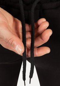 Czarne sznurki ze stoperami trzymane między palcami, na tle czarnej tkaniny. Sznurki mają teksturowany splot.