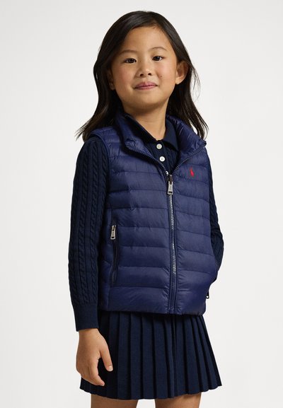 Marineblå vattert vest med høy krage, glidelåslukking foran og sidelengs lommer, kombinert med et marineblått plisséskjørt. Strukturerte ermer gir ekstra detaljer.