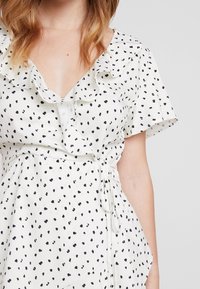 Valkoinen pusero, jossa on mustia pilkkuja, frillattuja kaula-aukkoja, lyhyet hihat ja vyötäisti. Kevyt kangas, jossa on löysä istuvuus.