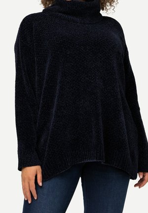 Personne portant un pull à col roulé en chenille bleu marine foncé ample et un jean bleu, debout devant un fond clair uni.