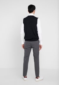 Schwarzer ärmelloser Pullover über einem weißen Hemd mit Kragen, kombiniert mit grauen Anzughosen und weißen Sneakern mit roten Akzenten.