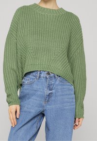 Personne portant un pull vert ample en maille, rentré dans un jean denim bleu clair taille haute, les mains reposant sur les côtés.