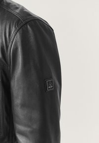 Manche de veste en cuir noir avec un patch de marque cousu présentant un logo trident et l'année 1982 sur un fond clair.
