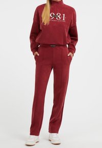 Bordowy dres składający się z bluzy z wysokim kołnierzem z logo oraz dopasowanych spodni zakończonych ściągaczem z elastycznym paskiem i kieszeniami bocznymi.