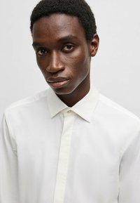 Camicia bianca con bottoni, dal tessuto liscio, con colletto a punta e pattina frontale. Il tessuto sembra leggero e fresco.
