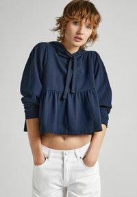Sudadera corta de color azul marino en tela suave con capucha con cordones, corte holgado y diseño peplum con puños acanalados. Combinada con pantalones blancos.