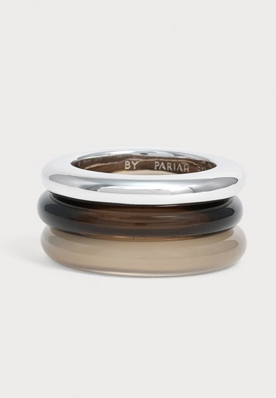 Trois anneaux lisses empilés avec des bords arrondis en argent, noir et beige sur un fond clair uni.