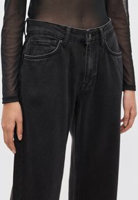 Čierne rifle z denimu s voľným strihom, vybavené piatimi vreckami, pútkami na opasok a viditeľným zipsom. Tkanina má hladký, matný povrch.