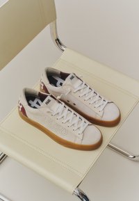 Baskets blanches et bordeaux avec semelles en gomme placées sur une chaise en cuir crème avec un cadre chromé.