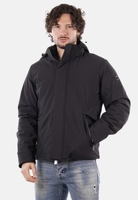 Uomo con capelli ricci, che indossa una giacca invernale nera con cappuccio e jeans blu sbiaditi, in piedi con una mano in tasca.