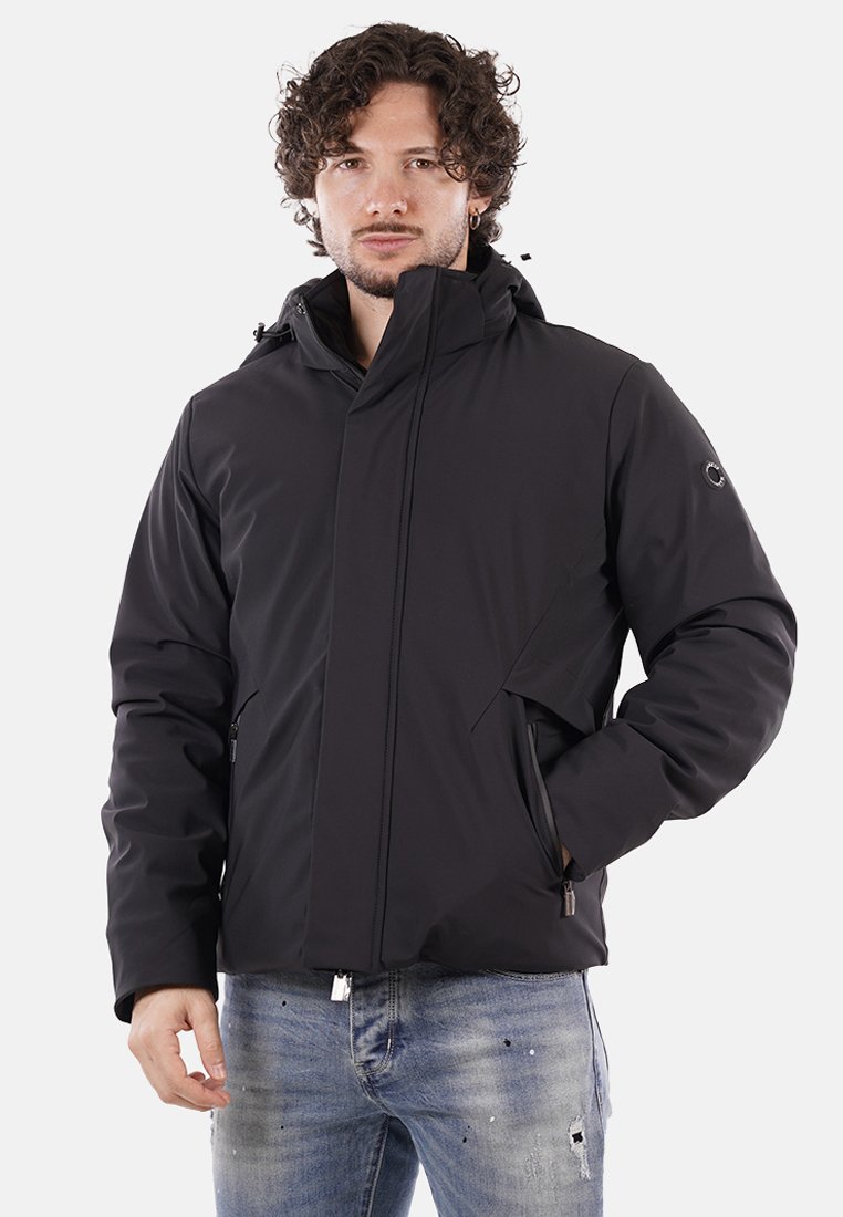 Uomo con capelli ricci, che indossa una giacca invernale nera con cappuccio e jeans blu sbiaditi, in piedi con una mano in tasca.