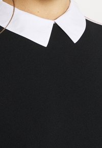 Schwarzer Strickpullover mit weißem Kragen. Der Kragen hat eine abgerundete Form, und der Stoff ist glatt mit feiner Textur. Keine weiteren Details sichtbar.