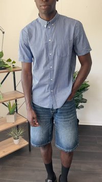 Camicia a maniche corte a righe blu e bianche con colletto abbottonato, abbinata a pantaloni corti di denim, calze nere e sandali a scivolo.
