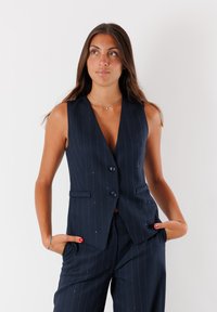 Gilet a righe blu navy con scollo a V profondo, due tasche frontali e due bottoni; realizzato in tessuto su misura con una texture liscia.