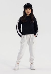 Ragazzina che indossa un berretto nero, una felpa nera con strisce bianche sui maniche, pantaloni cargo bianchi e sneakers bianche, in piedi con le mani in tasca.