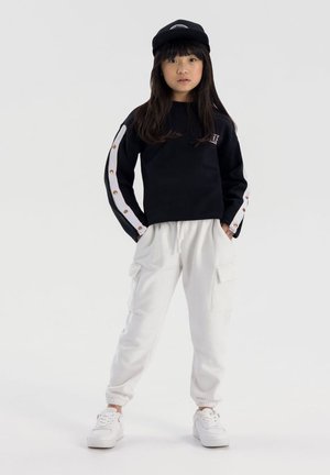 Ragazzina che indossa un berretto nero, una felpa nera con strisce bianche sui maniche, pantaloni cargo bianchi e sneakers bianche, in piedi con le mani in tasca.