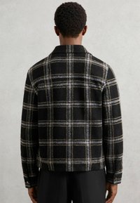 Sort og grå ternet jakke med en uldlignende tekstur, med spidse kraver og detaljer med knapmanchetter, set bagfra.
