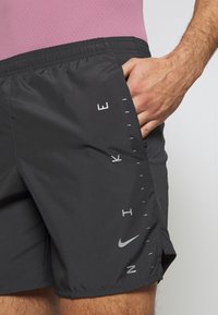 Nike miesten shortsit, mustat, kevyet materiaalit, joustava vyötäröbaari, harmaa logo ja pystysuuntainen teksti vasemmalla puolella.
