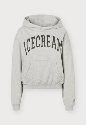 Le modèle porte un sweat à capuche gris ICECREAM, un short noir brodé et de hautes bottes noires sur un fond clair uni.
