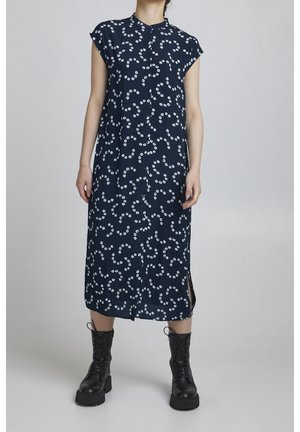 Vrouw in een marineblauwe midi-jurk met witte bloemenprint en kapmouwen, gecombineerd met zwarte veterlaarzen in combatstijl.