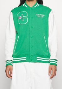 Chaqueta varsity verde y blanca con un parche de "C", cierre de botones, cuello y puños acanalados, y bolsillos laterales. Textura de tela suave.