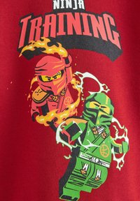 T-shirt rouge avec deux ninjas cartoon en action. L'un est rouge avec des flammes, l'autre est vert avec des effets de foudre. Le texte dit "Entraînement de Ninja".