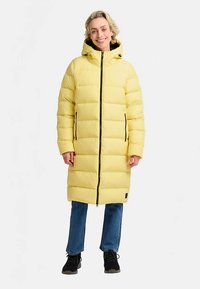 Lange gele pufferjas met capuchon, voorzien van een ritssluiting aan de voorkant, horizontale doorgestikte patronen en zijzakken. Draagt blauwe spijkerbroek en zwarte schoenen.