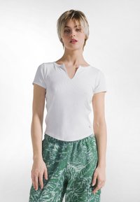 Bílý žebrovaný top s krátkými rukávy, výstřihem do V a zakřiveným lemem, kombinovaný se zelenými kalhotami s listovým vzorem.