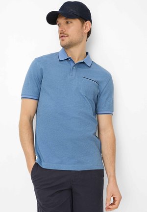 Mann trägt blaues Poloshirt mit Brusttasche und navyblaue Shorts, schaut seitlich, trägt schwarze Netzmütze, steht vor weißem Hintergrund.