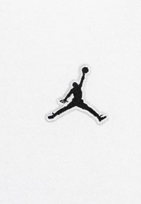 Logo Jumpman bordado en negro sobre tela blanca, que presenta la silueta de una persona saltando con un balón de baloncesto. Textura suave, diseño deportivo.