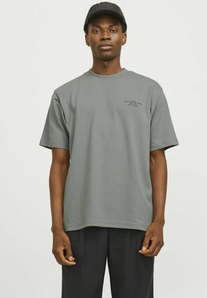 Graues Baumwoll-T-Shirt mit kurzen Ärmeln, Rundhalsausschnitt und dezentem Textdruck auf der Brust. Kombiniert mit einer dunklen Hose und einer schwarzen Kappe.