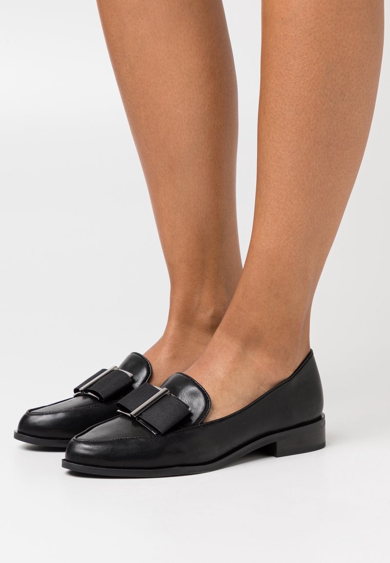 Mocassins plats en cuir noir avec de larges nœuds en tissu et des accents argentés, portés pieds nus sur fond blanc.
