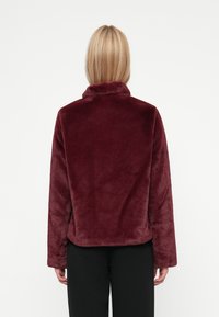 Chaqueta de pelaje sintético burdeos con cuello alto, mangas largas y una textura suave y esponjosa. Presenta un corte holgado y sin herrajes visibles.