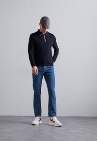 Μαύρο πλεκτό πουκάμισο με μακριά μανίκια και φερμουάρ στο λαιμό, μπλε τζιν και λευκά αθλητικά παπούτσια με καφέ λεπτομέρειες. Καπέλο με κατακόρυφες ρίγες σε ήσυχες αποχρώσεις.