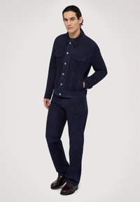 Marinblå mockajacka med bröstfickor, grå knappar och matchande mockabyxor. Modellen står med händerna i fickorna och har på sig bruna skor.