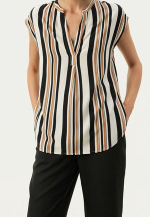 Sleeveless blouse with vertical black, white, and tan stripes, V-neck slit, worn with black pants, hands in pockets.